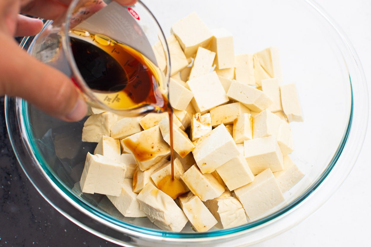 Pouring marinade over tofu cubes in glass bowl.