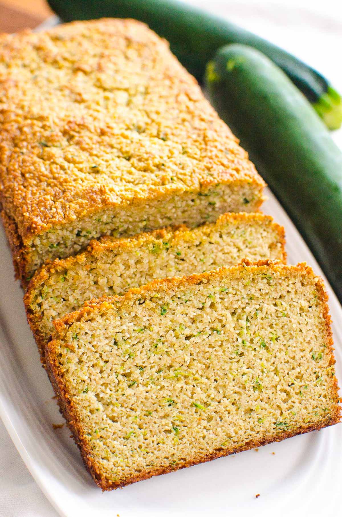 Sliced almond flour zucchini bread on a platter. More zucchini on a counter.