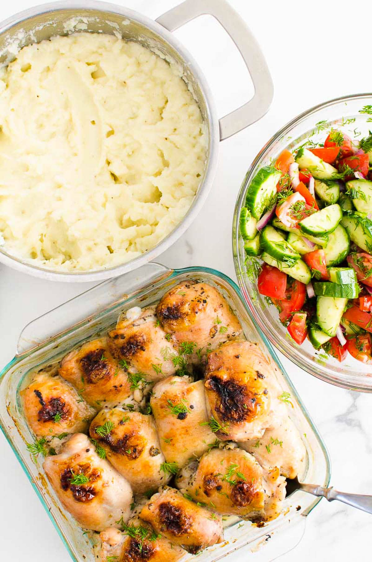 Oven baked chicken thighs, mashed potatoes in a pot and cucumber tomato salad in a bowl.