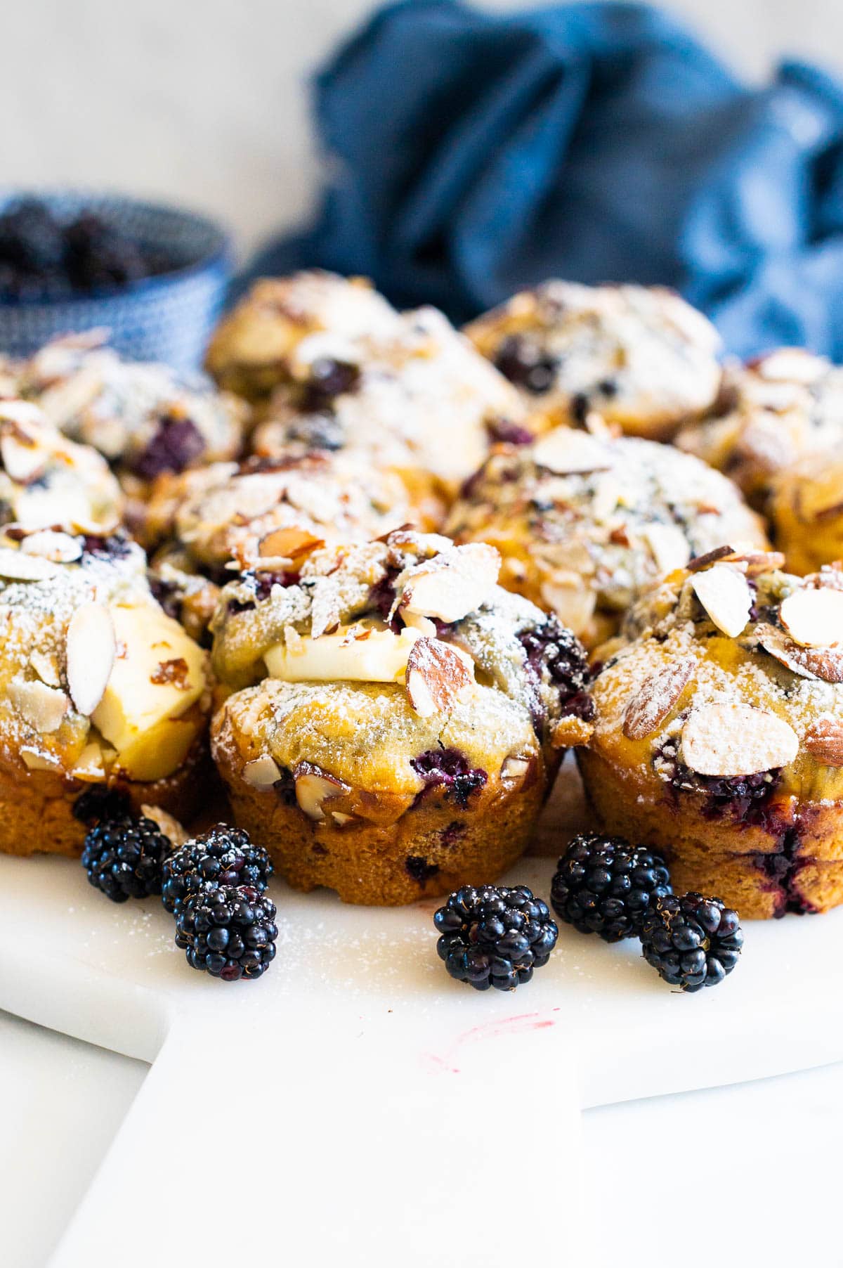 Blackberry muffins with cream cheese, sliced almonds and icing sugar served on a platter with fresh blackberries around them.