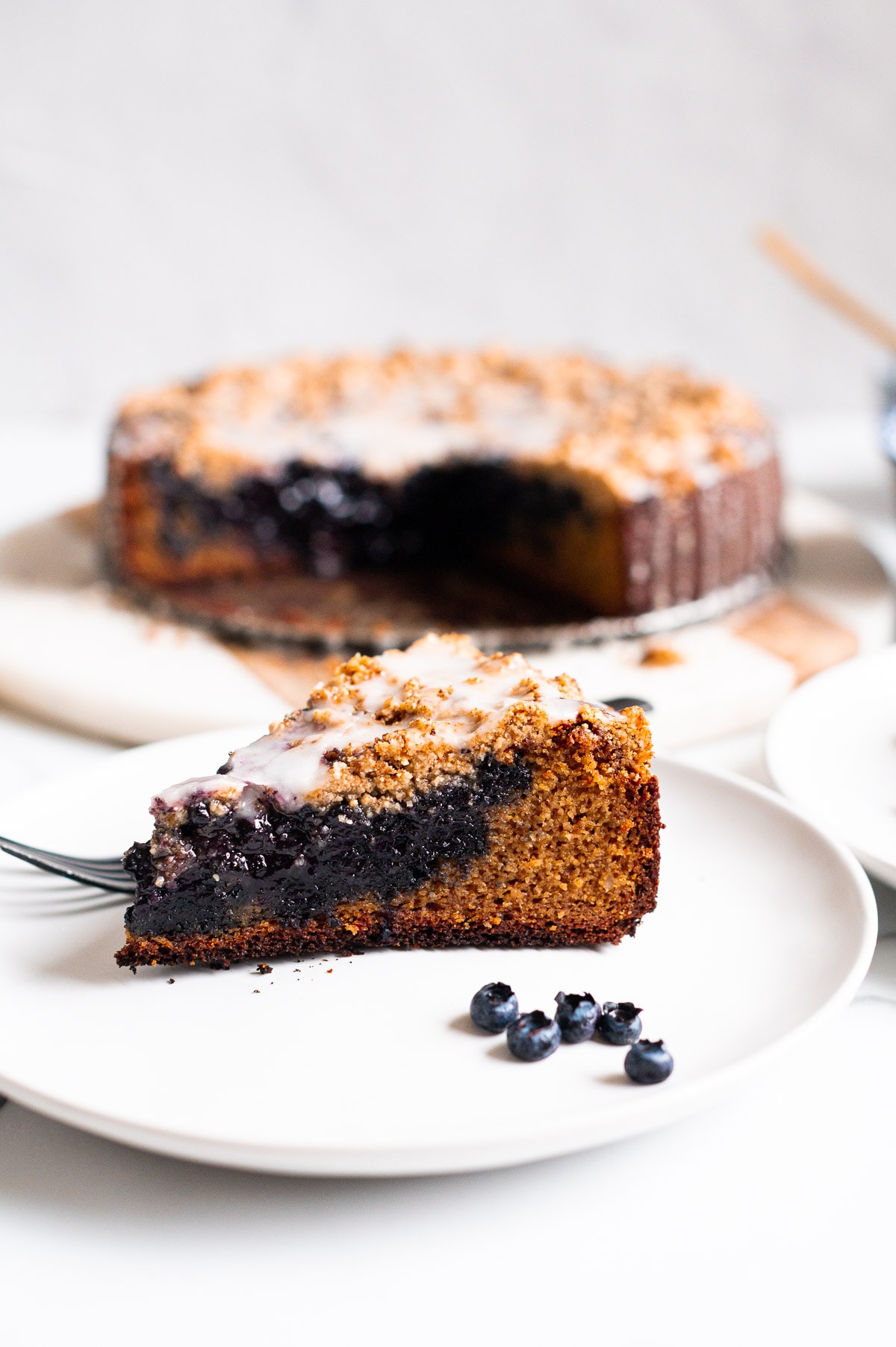 A slice of blueberry crumb cake with a glaze on a plate with blueberries and a fork. Cake in the background.