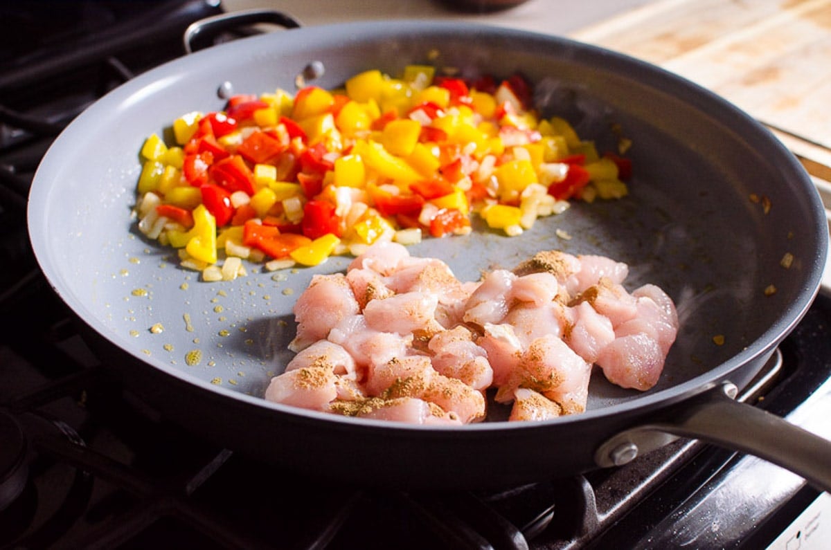 Sauteed veggies and cubed chicken breasts with seasonings in two piles in a skillet.
