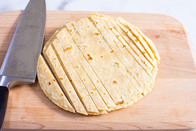 Sliced tortillas into thin strips on a cutting board with a knife.