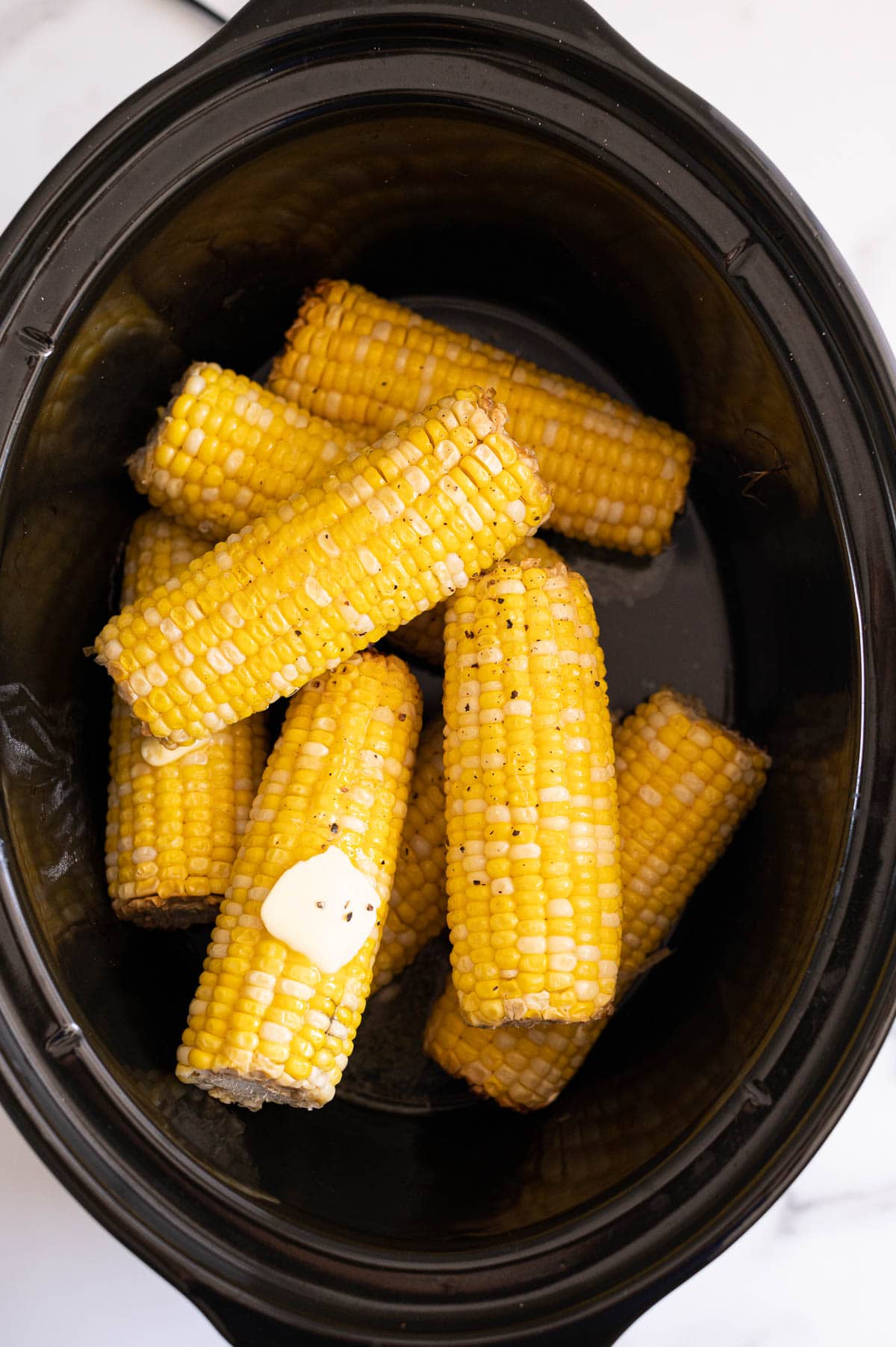 Crock pot corn on the cob smeared with butter and sprinkled with ground black pepper in a slow cooker insert.