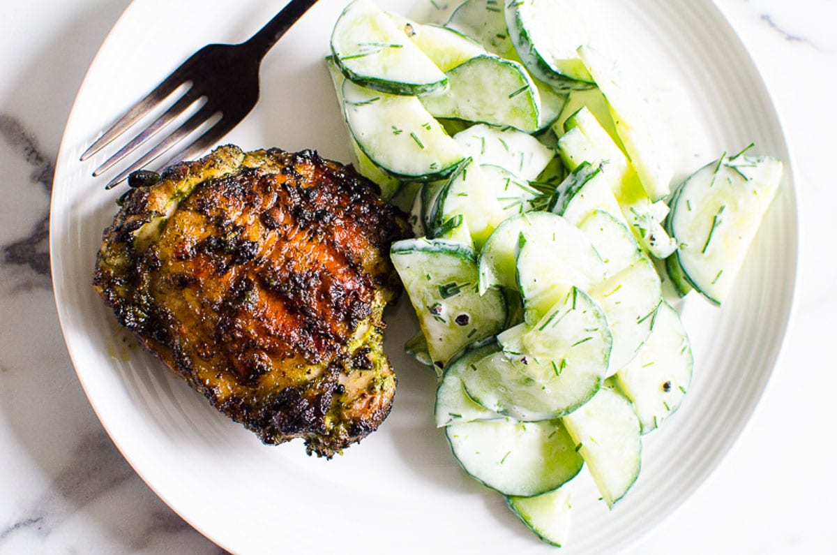 Grilled chicken thighs served with creamy cucumber salad on a plate with a fork.