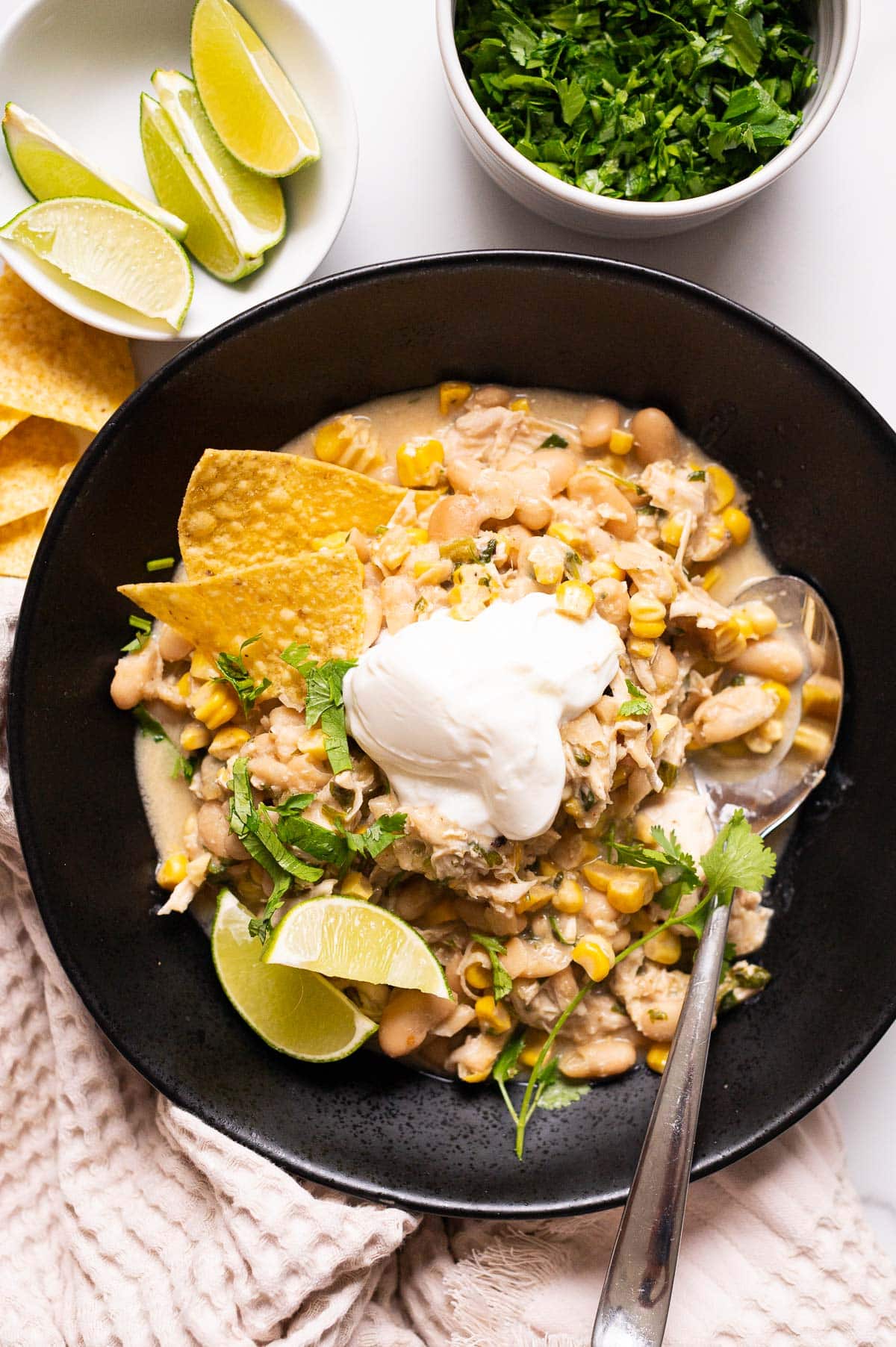 Crockpot white chicken chili served with yogurt, lime, cilantro and tortilla chips in black bowl with a spoon.