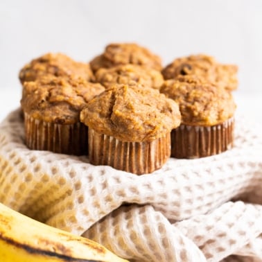 Seven banana carrot muffins on a linen towel and bananas nearby.