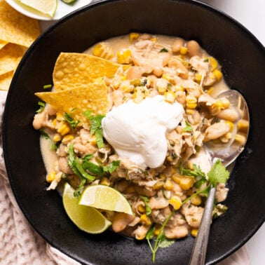 Crockpot white chicken chili served with yogurt, lime, cilantro and tortilla chips in black bowl with a spoon.