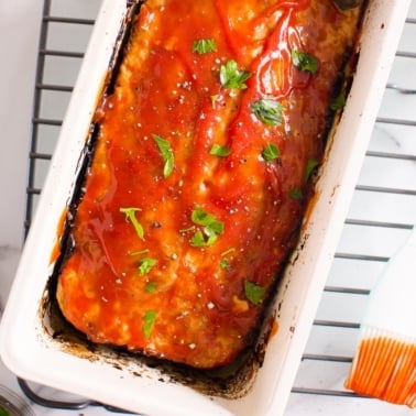 Turkey meatloaf with ketchup glaze in a pan on top of cooling rack with a brush.
