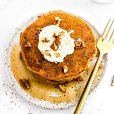 A stack of healthy pumpkin pancakes on a plate with maple syrup, pecans and yogurt.