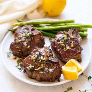 Three lamb loin chops served with asparagus and lemon slices on white plate.