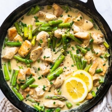 Lemon chicken and asparagus in creamy sauce with lemon slices in cast iron skillet.