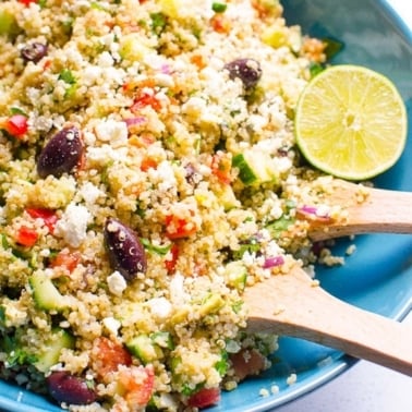 Mediterranean quinoa salad in a blue bowl with serving tongs.