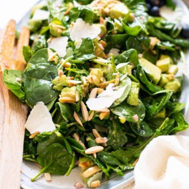 Spinach salad with avocado, almonds, parmesan cheese on a serving platter with wooden tongs.