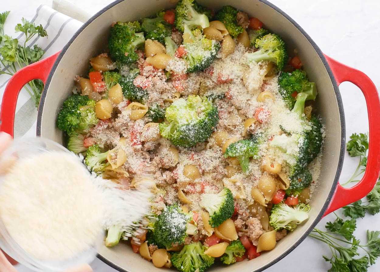 Parmesan cheese being poured into a pot with broccoli, pasta and ground turkey.