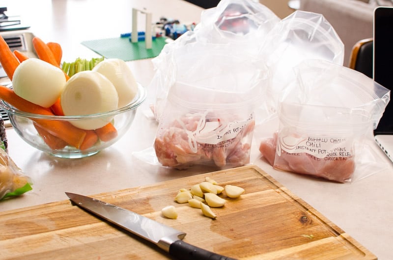 Assembled freezer meals in bags, vegetables in a bowl and garlic on a cutting board with a knife.