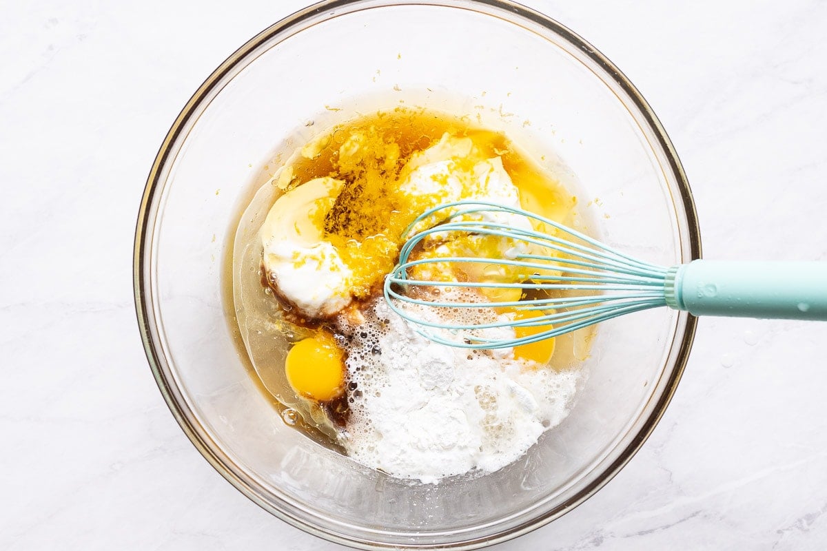 Eggs, yogurt, maple syrup, oil, lemon zest and juice, vanilla, and baking staples in bowl with a whisk.