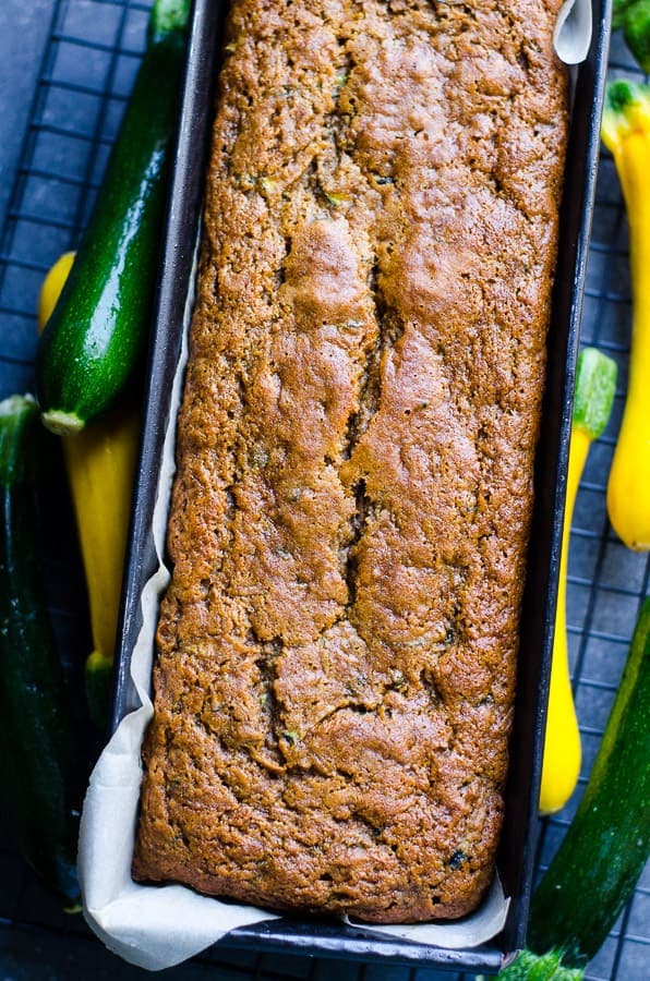 Healthy zucchini bread in a loaf pan on a metal rack surrounded by summer squash and zucchini.