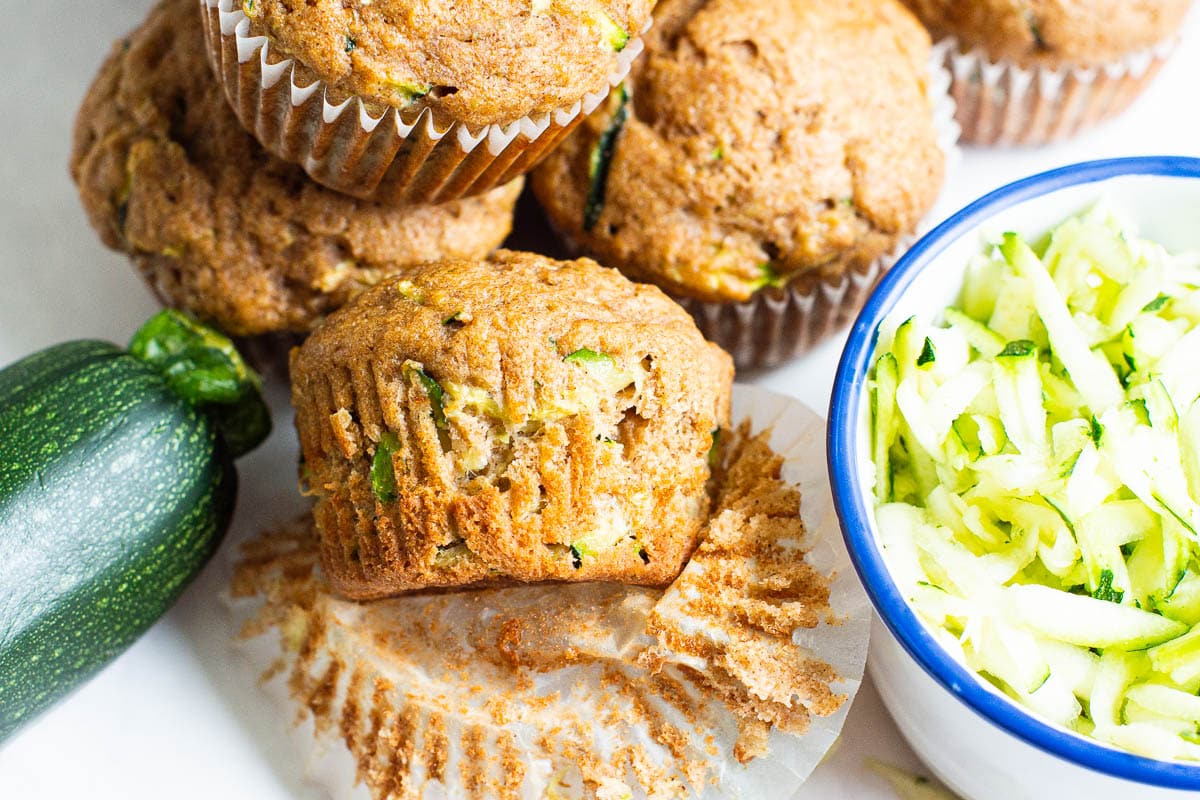 Healthy zucchini muffins with a bowl of shredded zucchini and one muffin unwrapped.