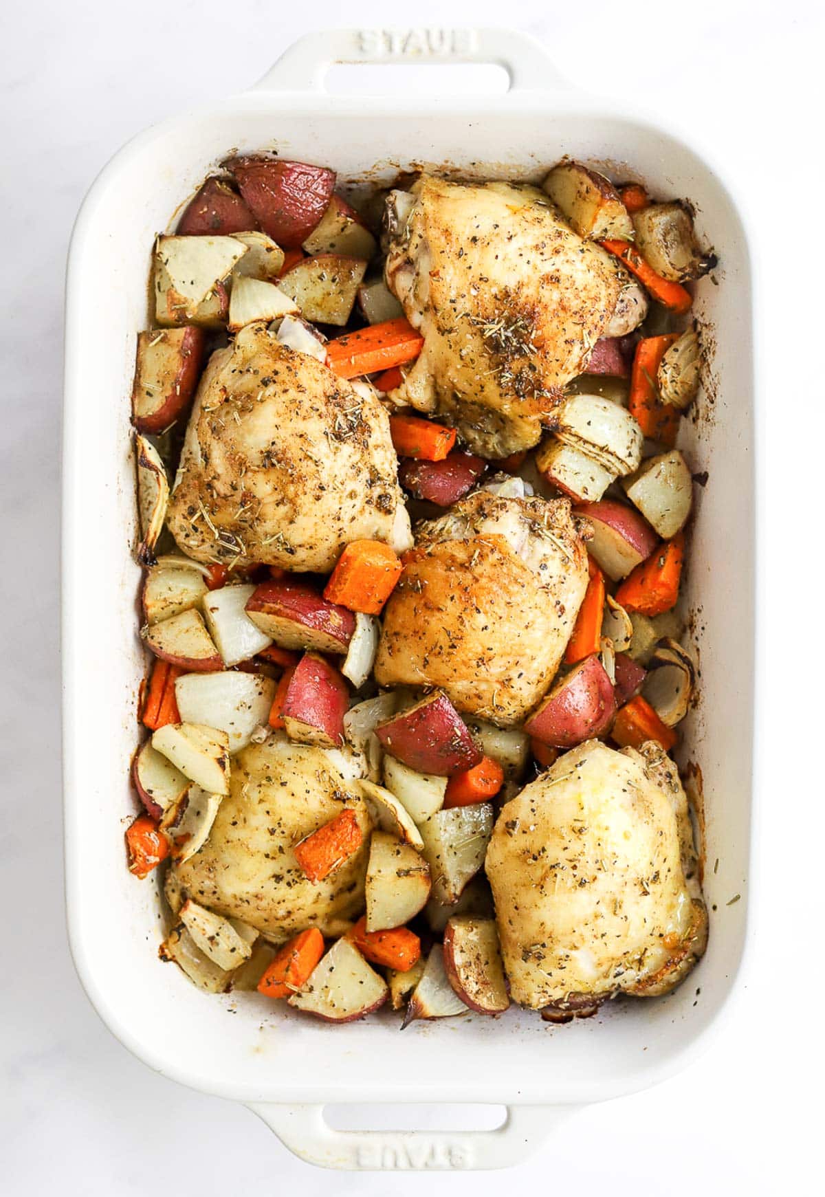 Roasted chicken thighs and potatoes and carrots in white baking dish.