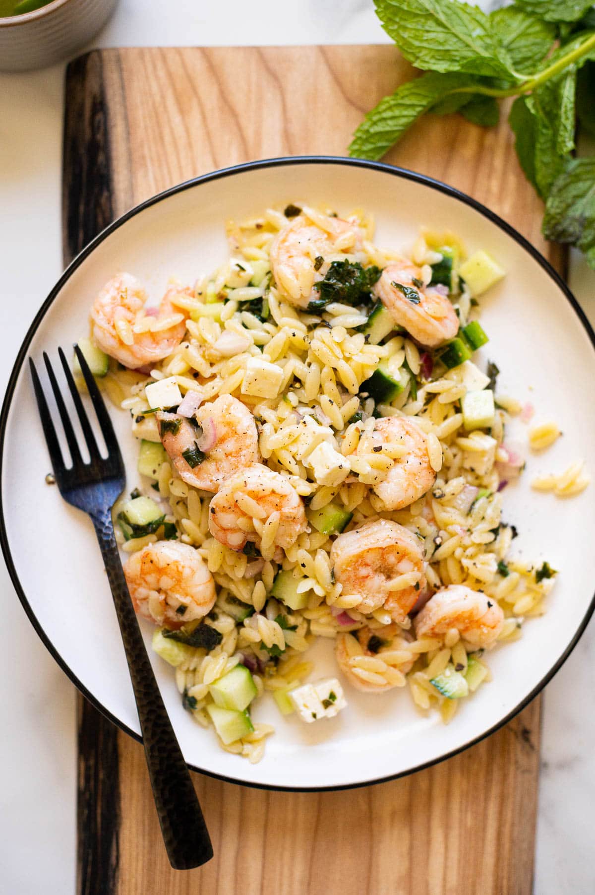 Ina Garten's shrimp orzo salad served on a plate with a fork on a wooden board. Mint as a garnish on a counter.