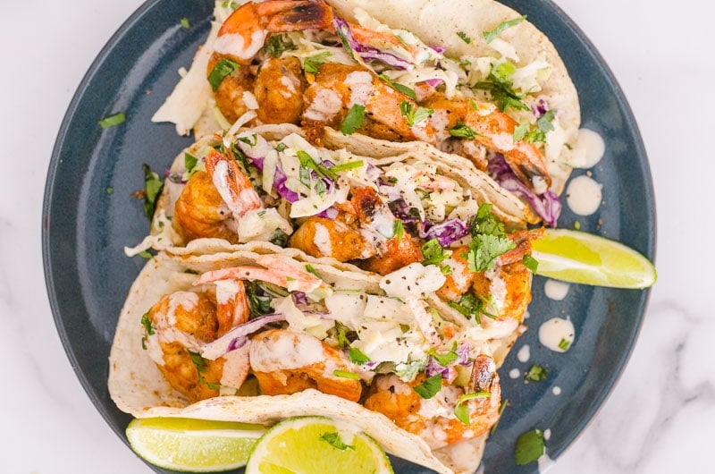 Three healthy shrimp tacos served with slaw and lime wedges on a plate.