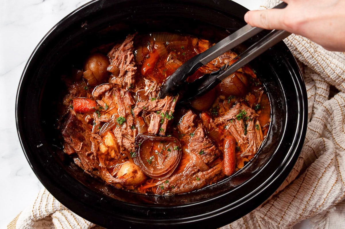Person holding tongs inserted into crock pot with shredded beef and veggies in au jus.