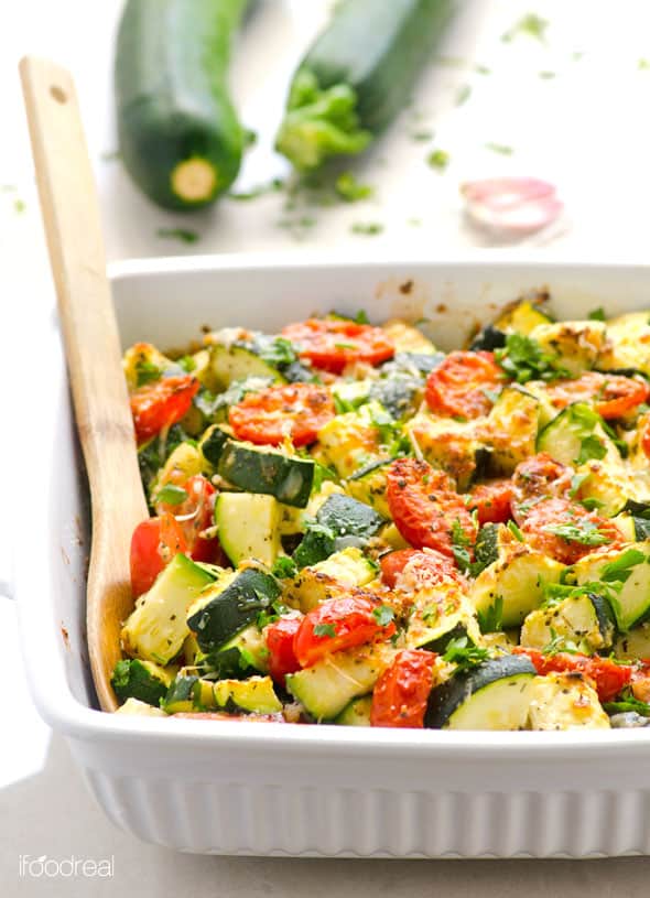 Zucchini tomato bake in white baking dish with wooden spoon and more zucchini and garlic in the background.