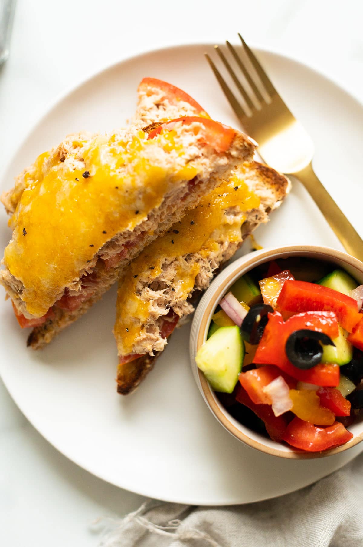 Tuna melt cut in half and served with tomato cucumber bell pepper salad on a plate.
