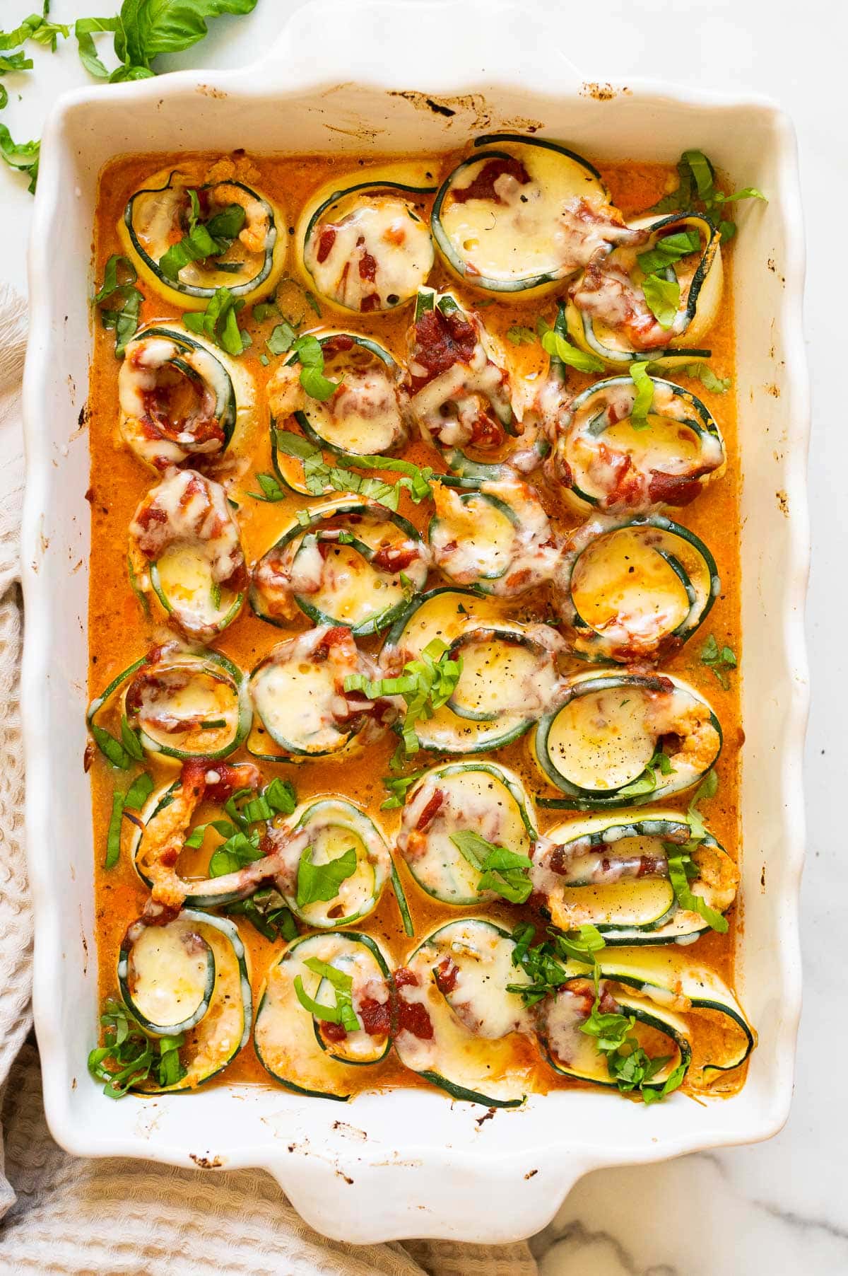 Zucchini lasagna roll-ups arranged in a baking pan with sauce and garnished with basil.