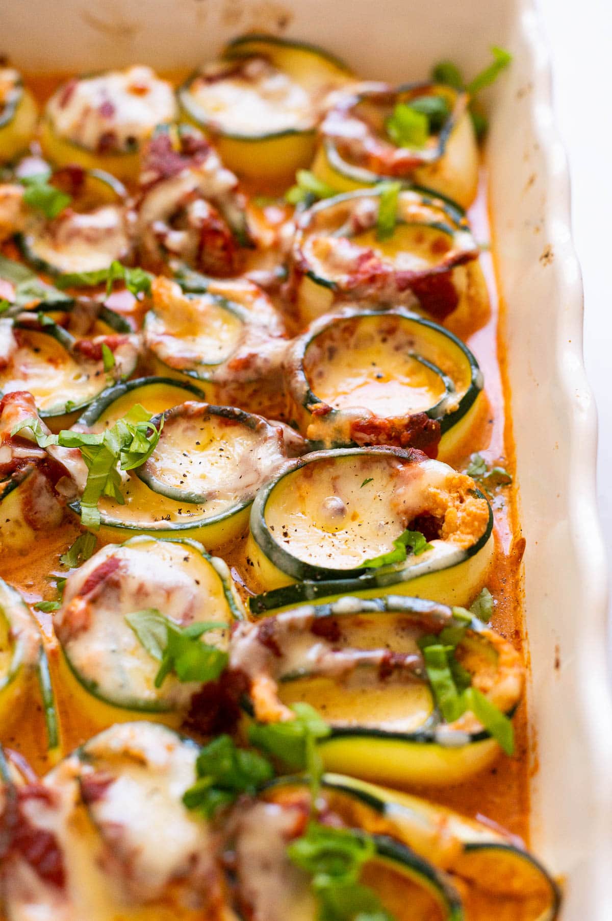 Side view of zucchini lasagna roll-ups in a baking dish with sauce.